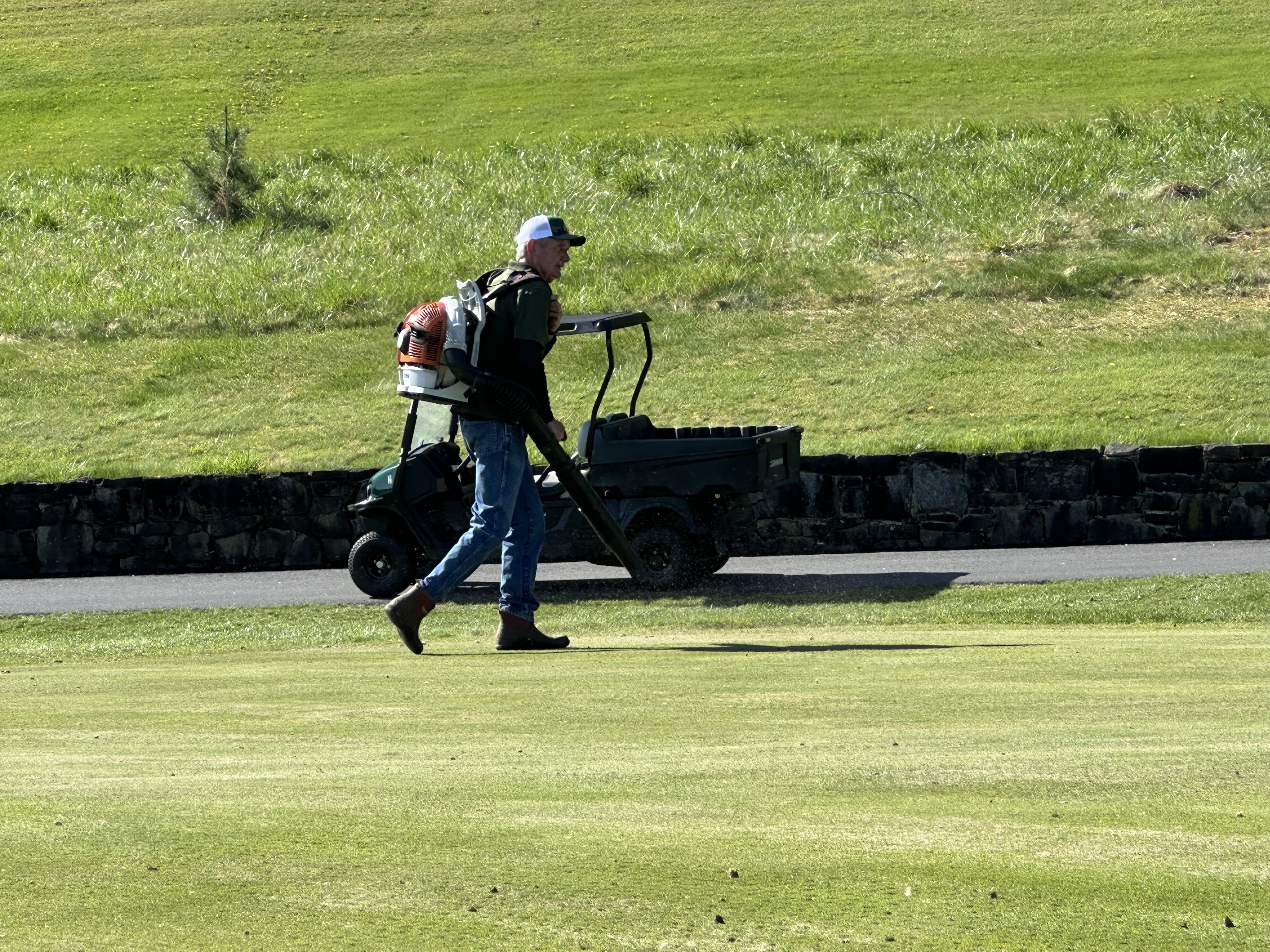 Brad carrying a backpack blower across a fairway with a utility vehicle in the background
